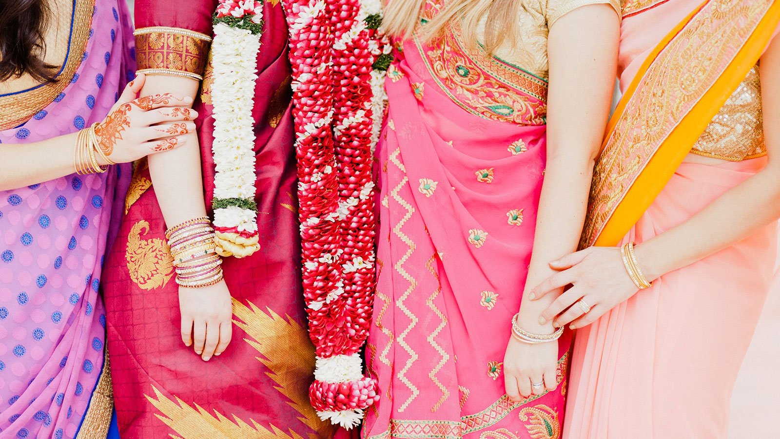 Women in colorful saris with henna, bangles, and floral garlands link arms together at a Miami wedding celebration